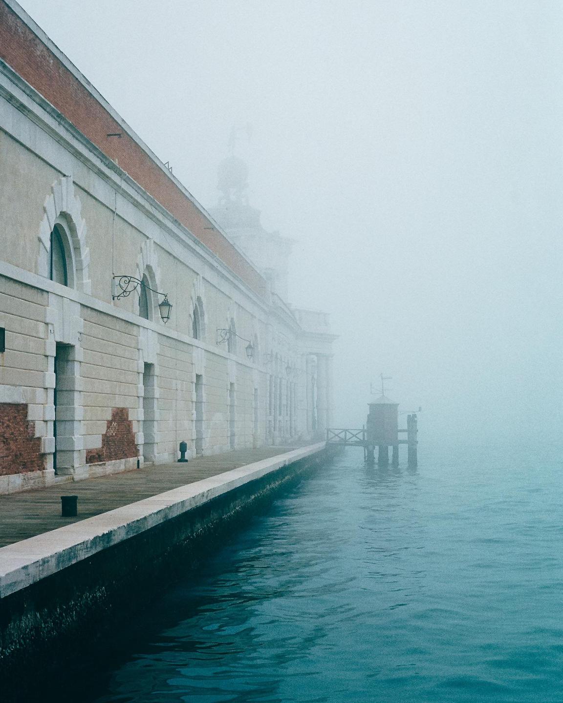 A building with arched windows stands beside a canal on a foggy day.