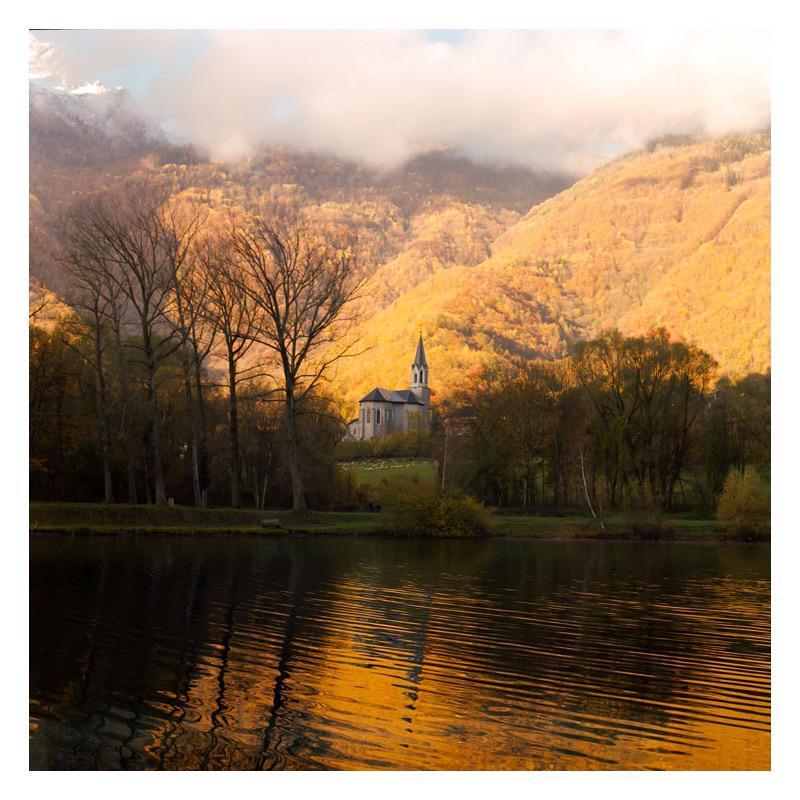 A church with a tall steeple sits on a hill overlooking a lake, surrounded by trees and mountains with autumn foliage.