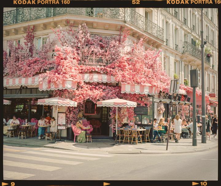 A cafe with pink awnings and pink flowers covering its facade has people seated at tables on the sidewalk.