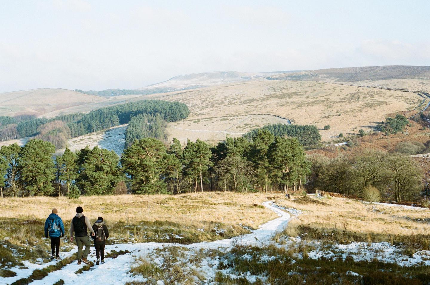 Three people walk on a snowy path through a winter landscape with hills and trees.