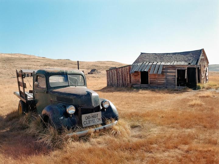 An old truck with a "DO NOT CLIMB ON" sign on its bumper sits in a field of dry grass next to a dilapidated wooden building.