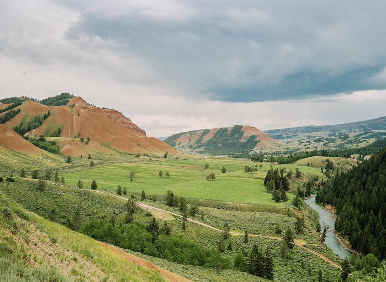 A wide valley with green fields, scattered trees, and a winding river is surrounded by hills and mountains under a cloudy sky.