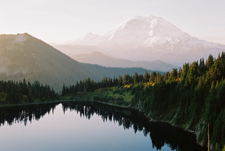 A snow-capped mountain rises above a forest and a lake reflecting the trees.