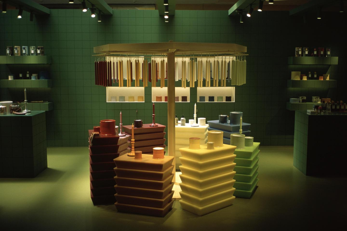 A store interior features candles and other products displayed on shelves and tables against a green tiled wall.