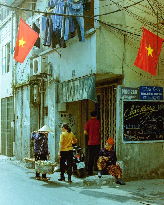 A street vendor with a conical hat carries baskets on a pole while other people stand and sit on a narrow street in Hanoi, Vietnam.