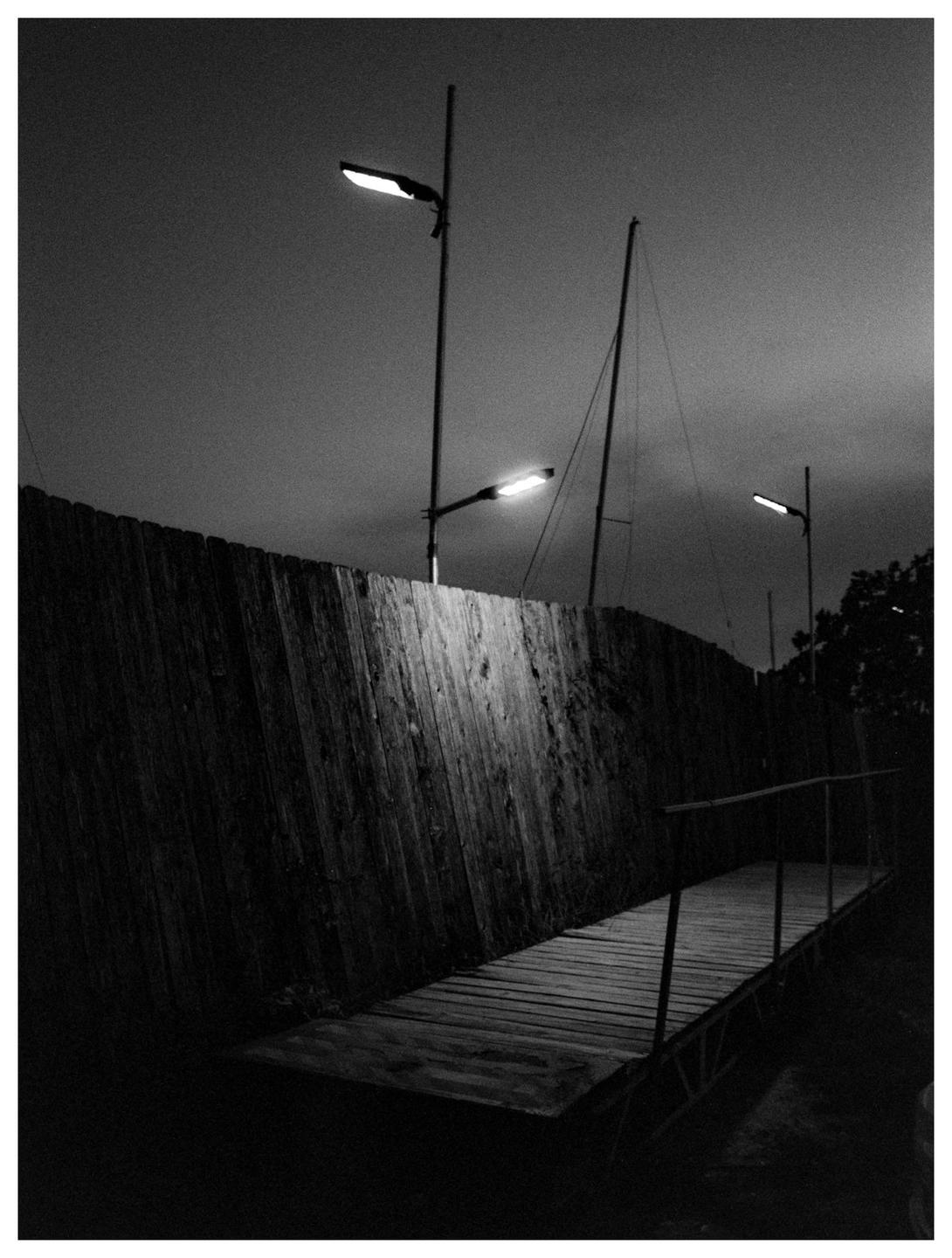 A wooden boardwalk and fence are illuminated by streetlights at night, with a sailboat mast visible in the background.
