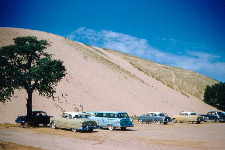 A group of vintage cars are parked at the base of a large sand dune with people climbing it.