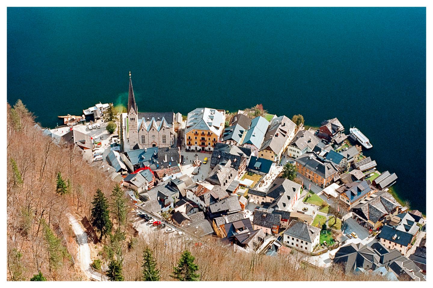 A village with a prominent church steeple is nestled on the shore of a large lake, surrounded by a forest of bare trees.