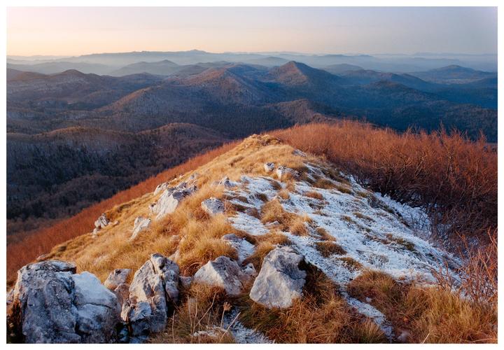 A mountain ridge covered in dry grass and patches of snow overlooks a valley of forested hills.