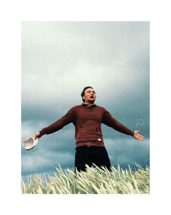 A man stands in a field of tall grass with his arms outstretched and mouth open, holding a hat in one hand, under a cloudy sky.