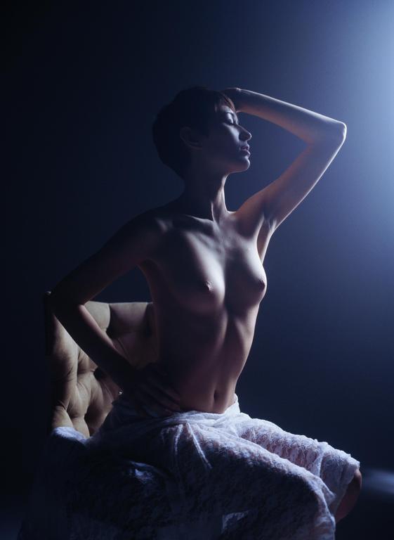 A woman with short hair sits on a chair, illuminated by a bright light from the right.