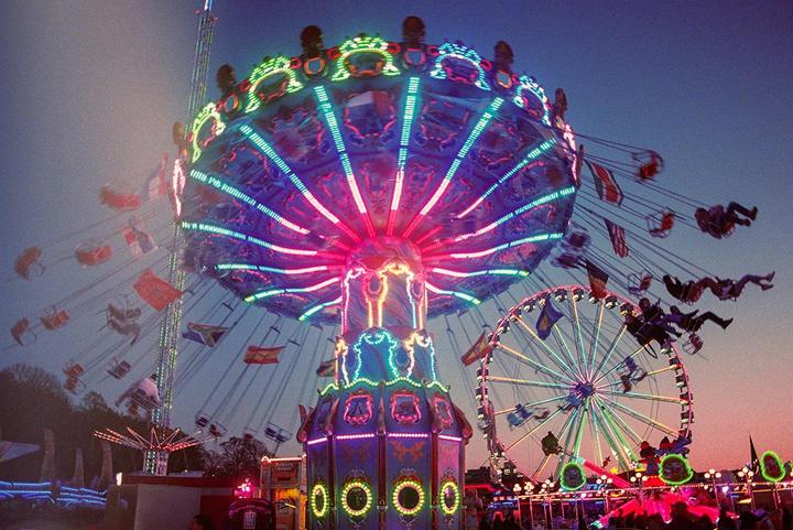 A brightly lit swing ride and ferris wheel are visible against a dusk sky at a fairground.