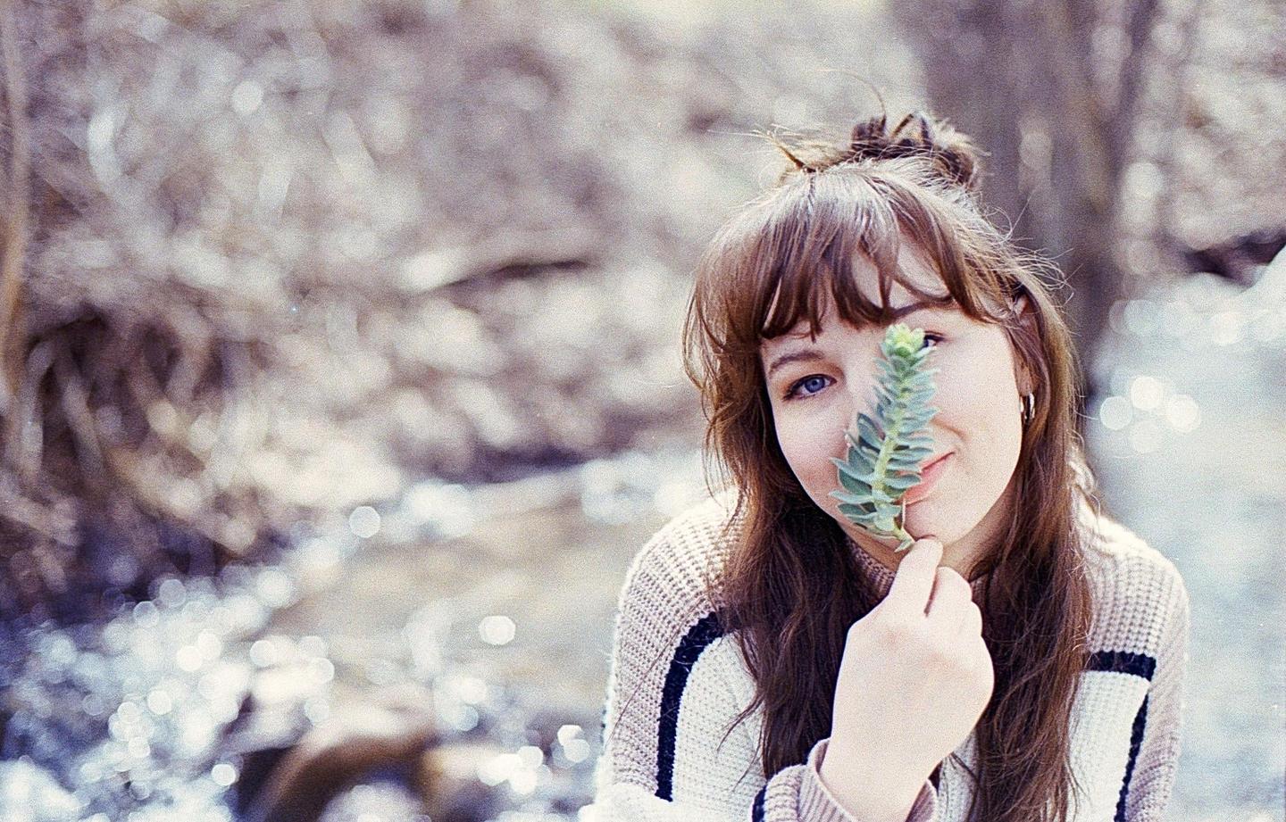 A woman with long brown hair holds a green plant in front of her face, partially obscuring one eye.