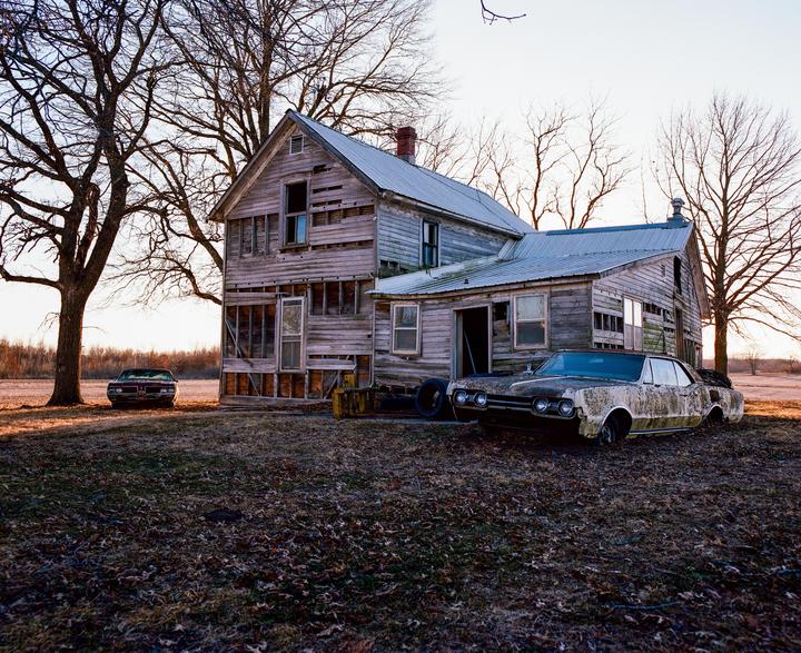 An abandoned wooden house stands next to two old cars in a field with bare trees under a clear sky.