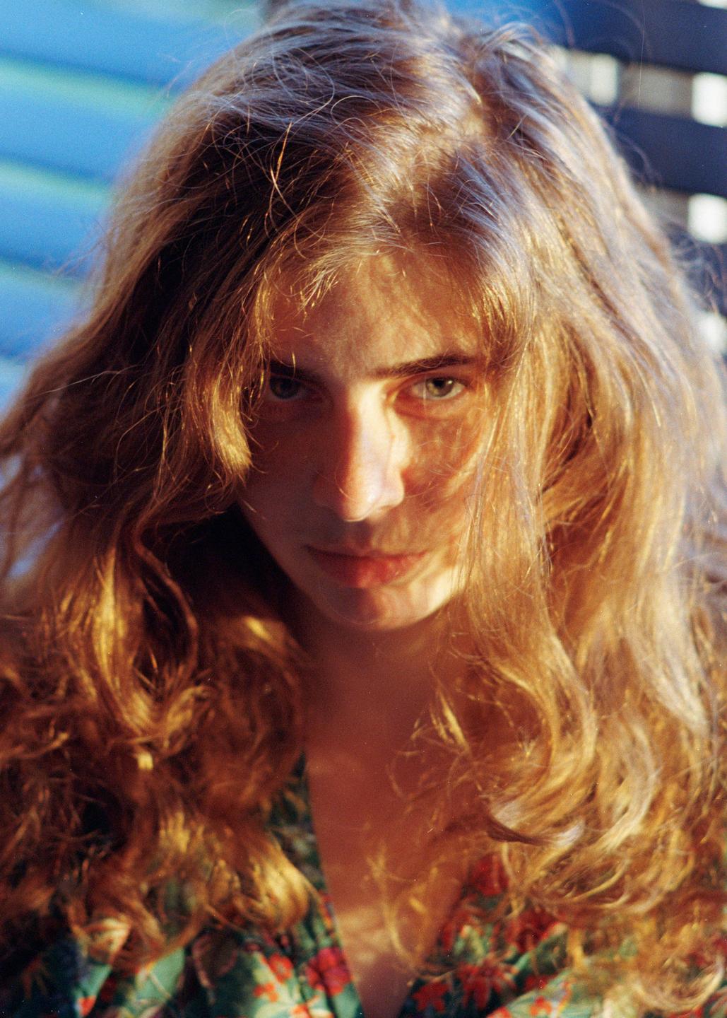 A woman with long, wavy hair looks directly at the viewer, with sunlight illuminating her hair and face.
