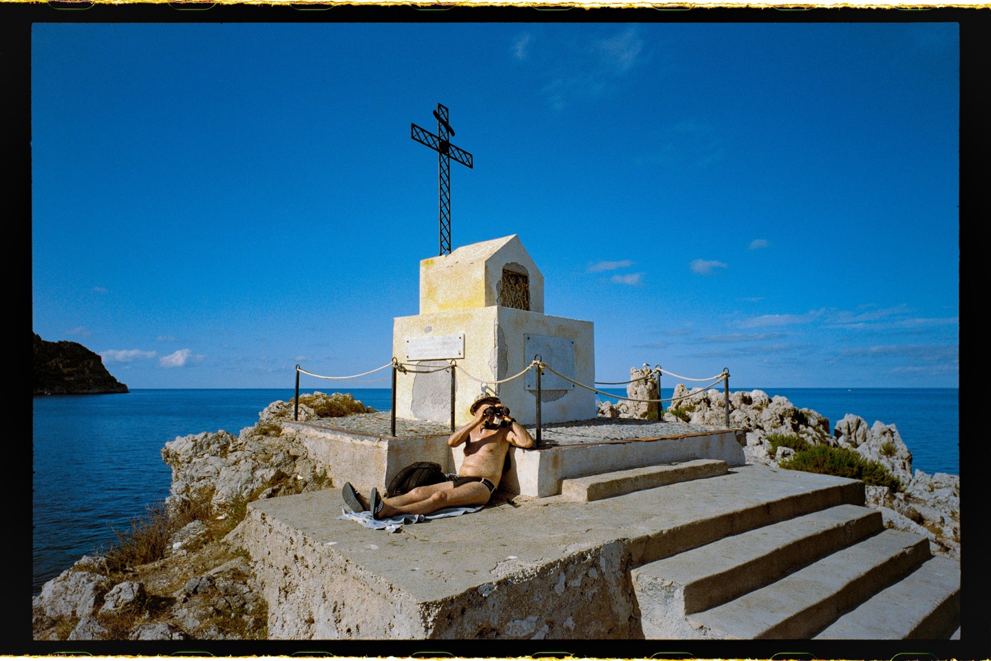 A man in a hat and swim briefs reclines on a stone platform next to a monument with a cross, looking through binoculars at the sea.