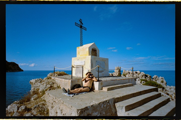 A man in a hat and swim briefs reclines on a stone platform next to a monument with a cross, looking through binoculars at the sea.