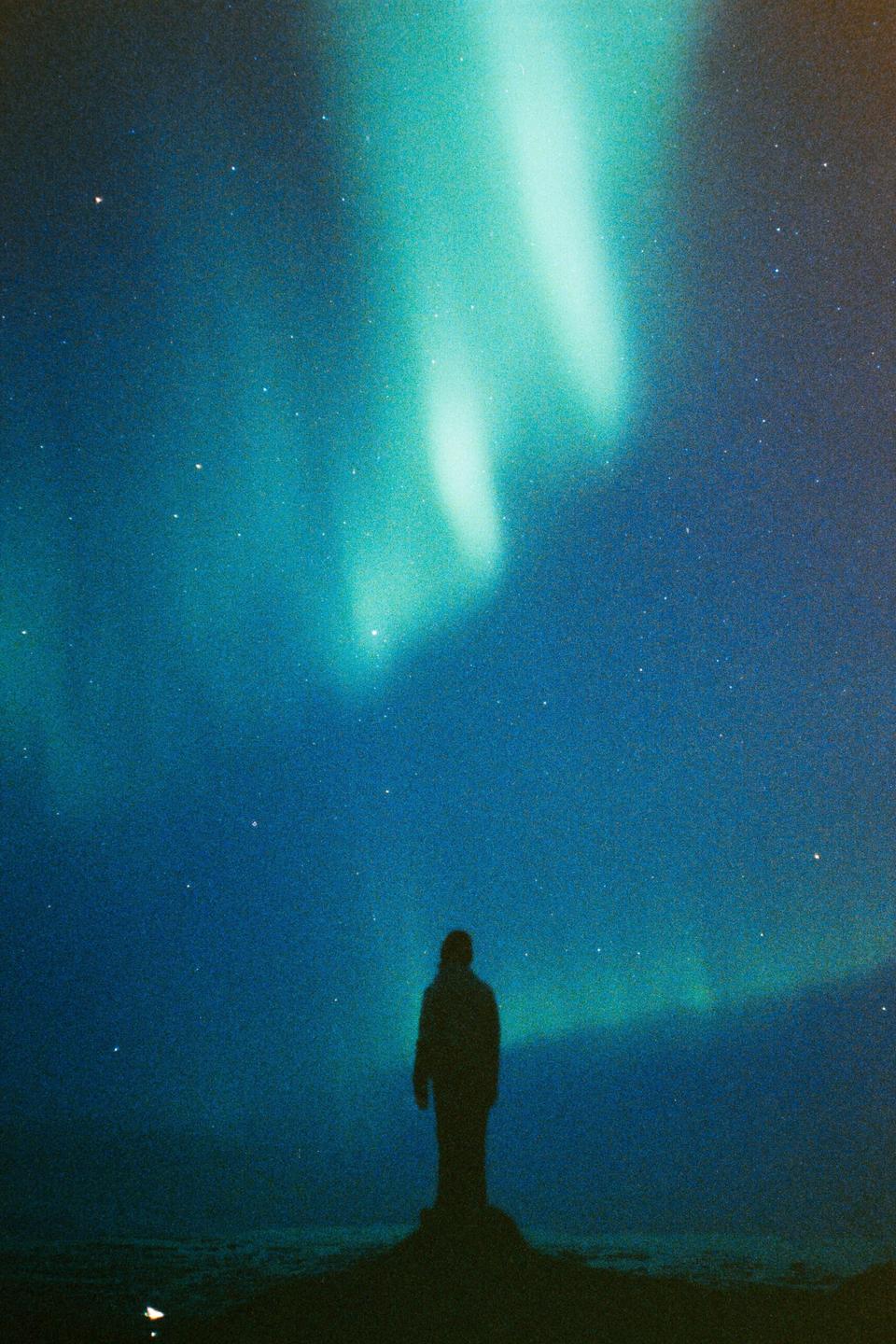 A lone figure stands silhouetted against a starry night sky illuminated by the aurora borealis.