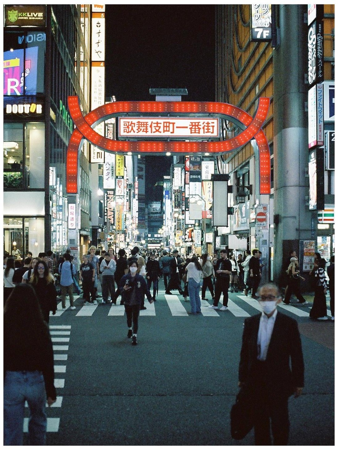 A busy street in Tokyo at night is filled with pedestrians crossing a crosswalk under a large red archway sign.