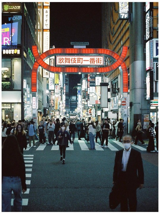 A busy street in Tokyo at night is filled with pedestrians crossing a crosswalk under a large red archway sign.