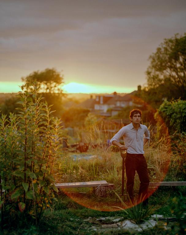A man stands in a garden with a shovel at sunset, with houses visible in the distance.