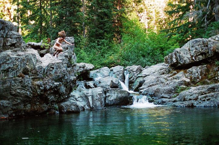 A person jumps from a rocky cliff into a natural pool with a waterfall in a forest.