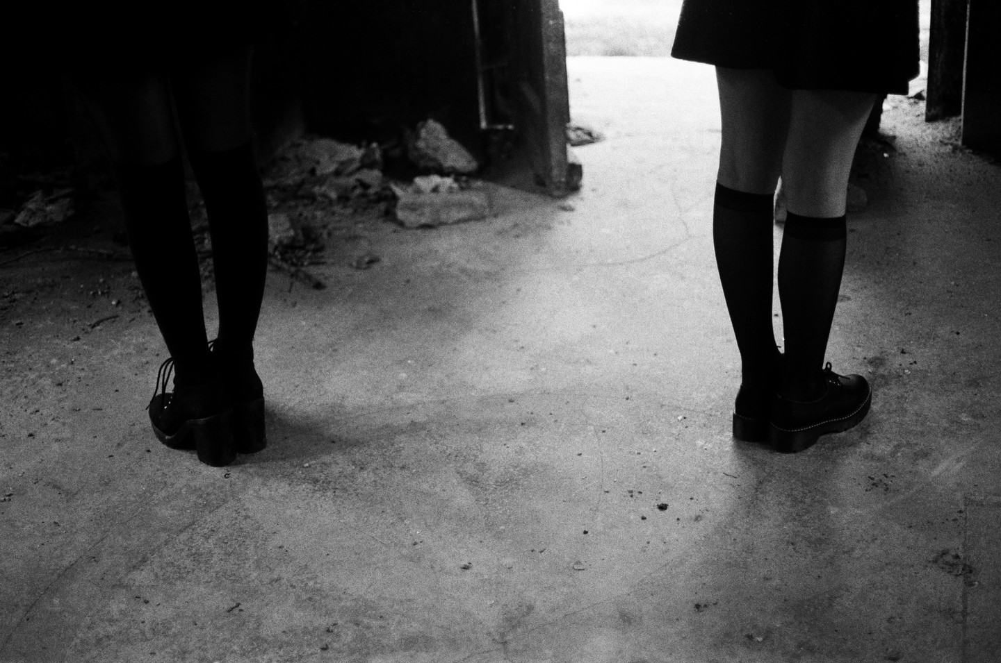 Two women stand on a concrete floor, one facing away and the other facing towards the viewer, with only their legs and feet visible.