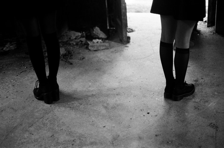 Two women stand on a concrete floor, one facing away and the other facing towards the viewer, with only their legs and feet visible.