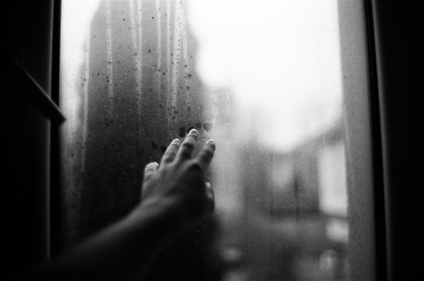 A hand presses against a dirty, streaky window, with a blurry, bright background.