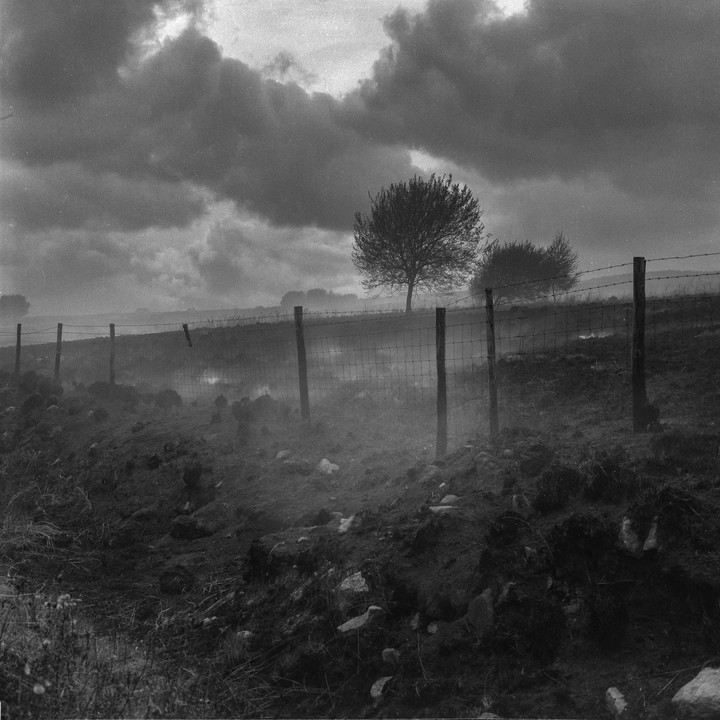 Smoke rises from a recently burned hillside with a fence and trees under a cloudy sky.