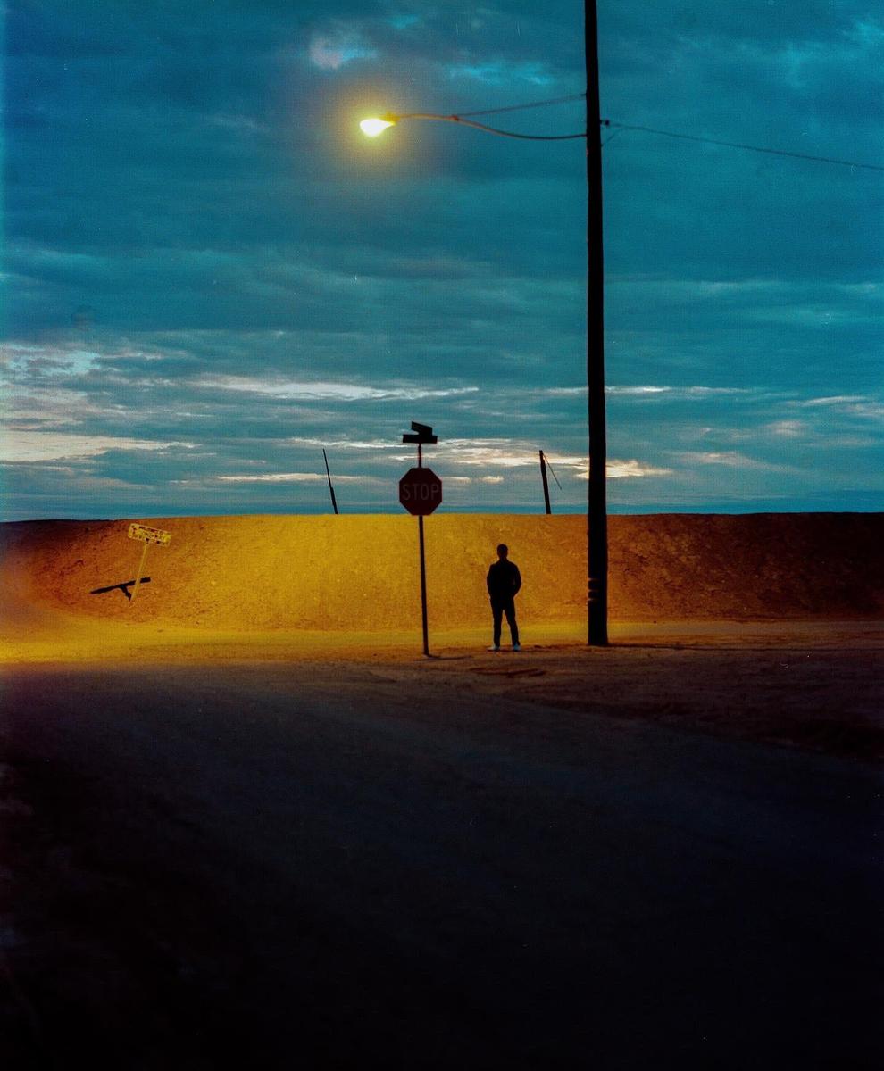 A man stands next to a stop sign and a utility pole under a street light at dusk.