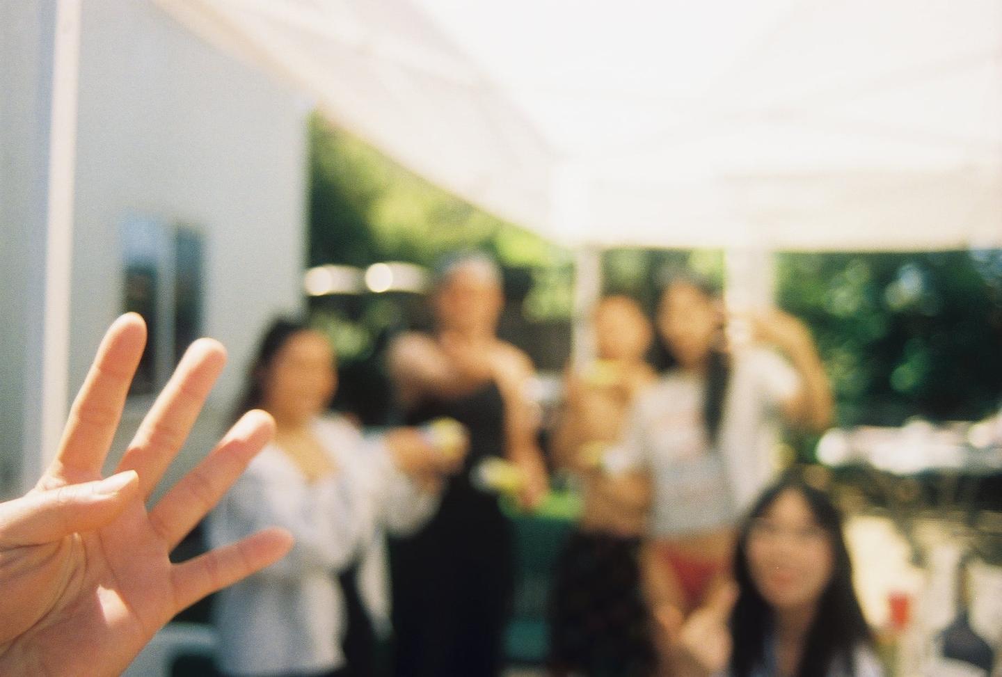 A hand with four fingers extended is in the foreground, while several blurry people are visible in the background under a white canopy.