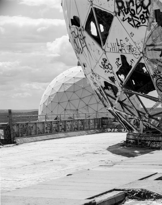 Two geodesic domes, one covered in graffiti and partially destroyed, stand on a concrete surface under a cloudy sky.