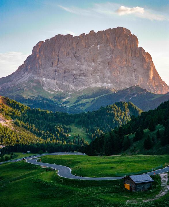 A winding road leads through green hills towards a large mountain under a bright sky.