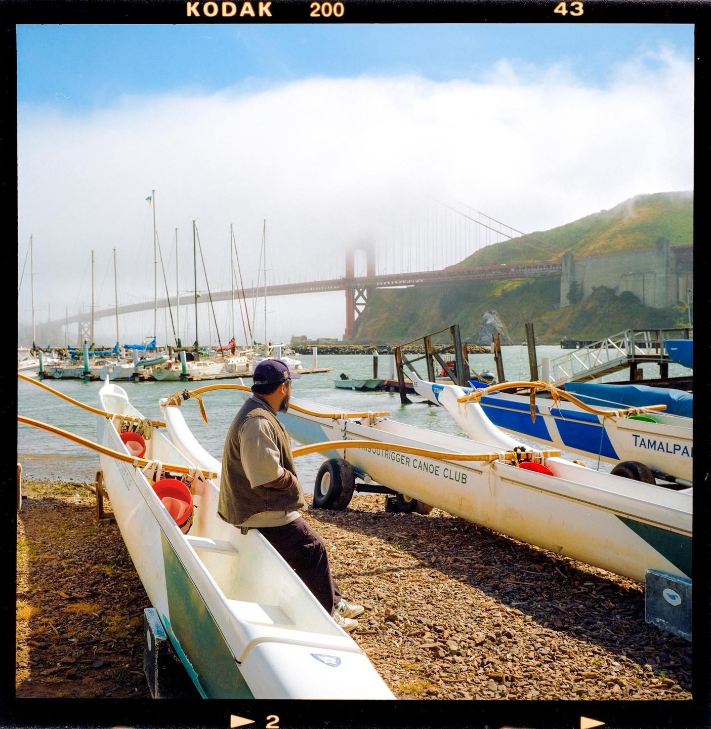 A man sits on a canoe on a rocky shore with a bridge and sailboats in the background.