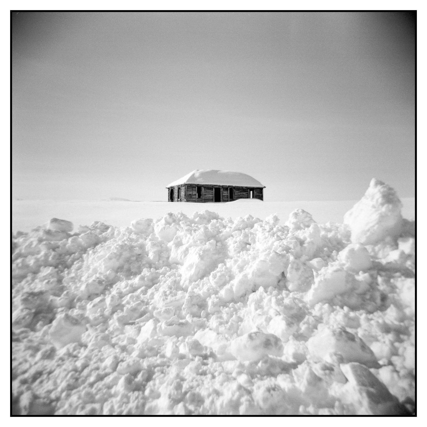 A small wooden cabin with a snow-covered roof is visible behind a large pile of snow.