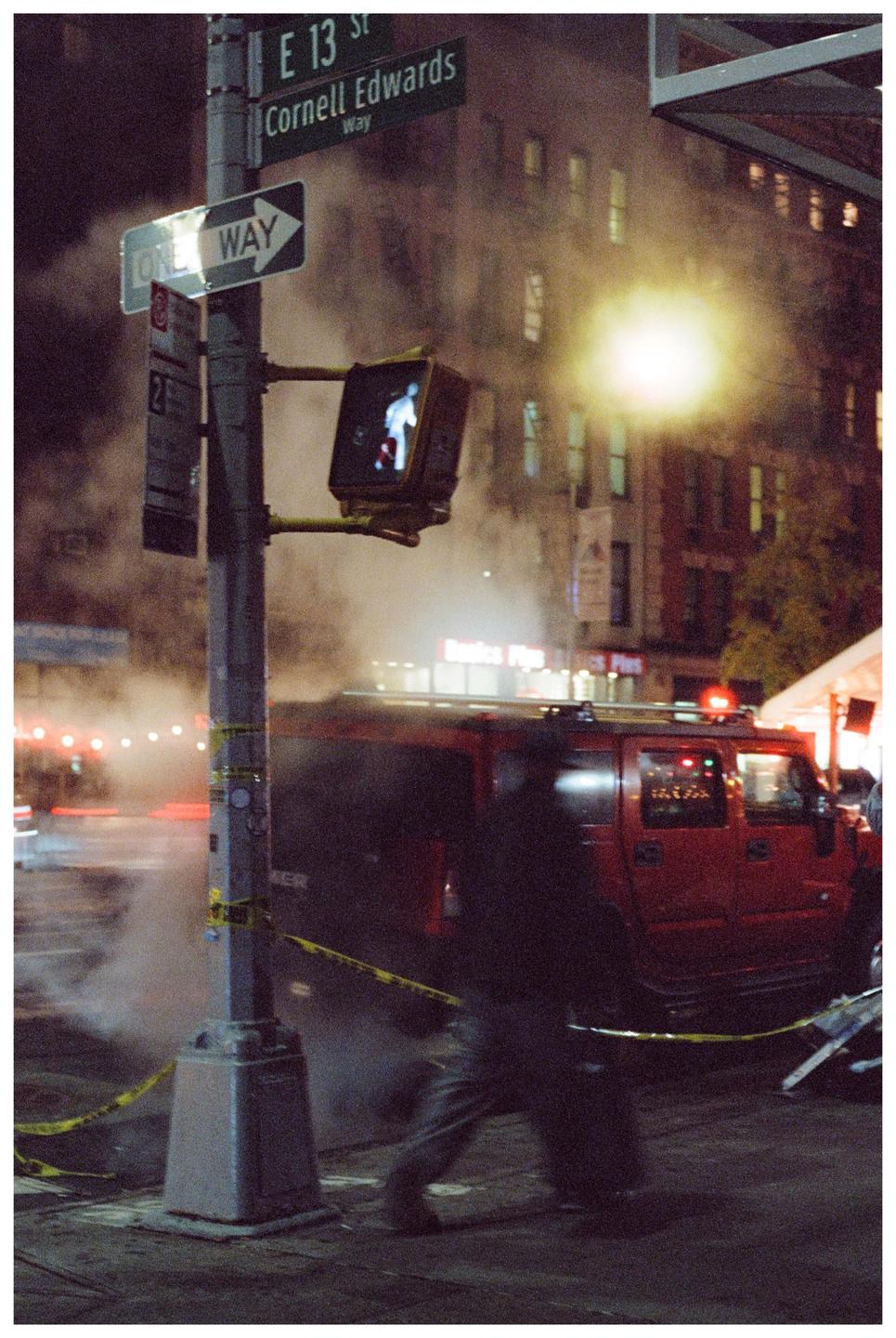 A street sign for Cornell Edwards Way and E 13 St is visible above a one way sign and a pedestrian crossing signal, with a red vehicle and a person in motion blur in the background.