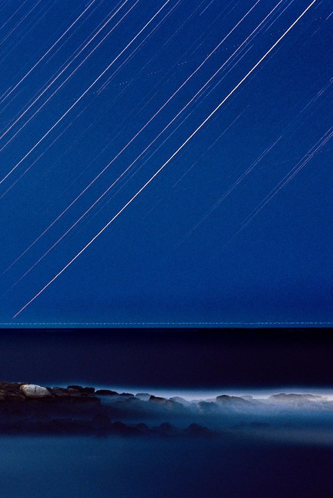 Star trails streak across a deep blue night sky above a rocky coastline with misty water.