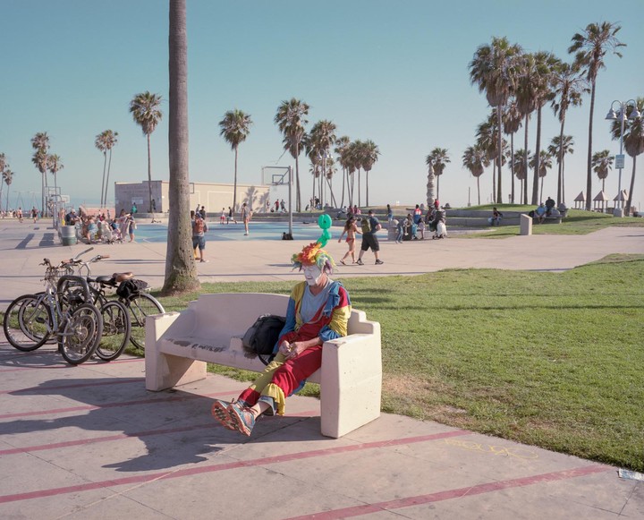 A clown in a colorful costume and wig sits on a bench next to bicycles, with palm trees and people in the background.