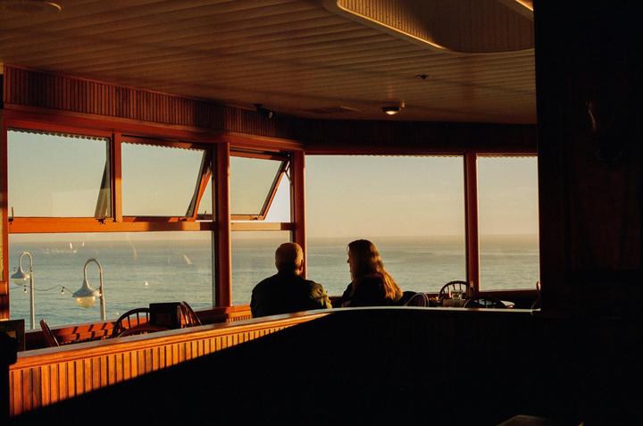 A man and a woman sit at a table in a restaurant with large windows overlooking the ocean at sunset.
