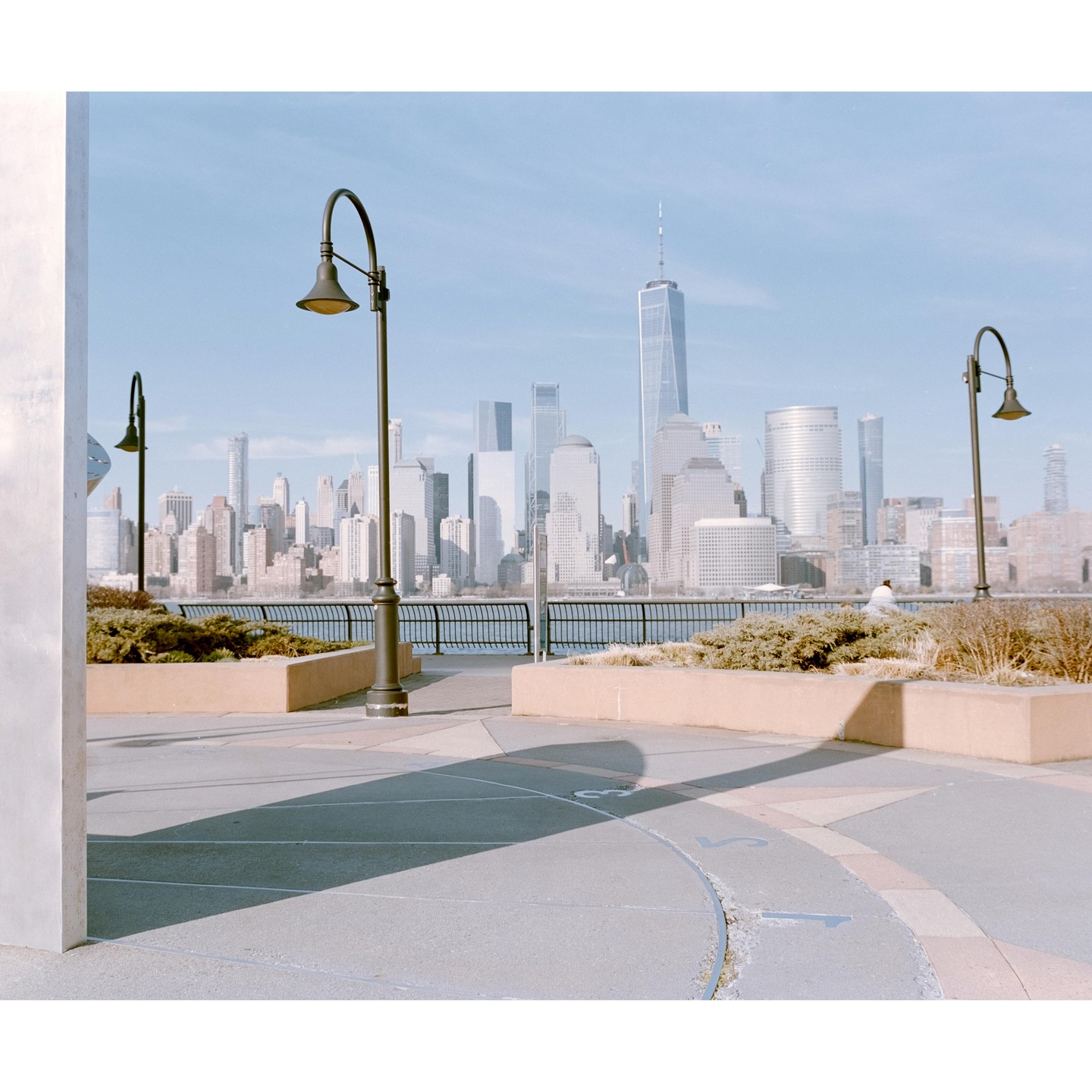 A waterfront promenade with streetlights and planters overlooks a city skyline across the water.