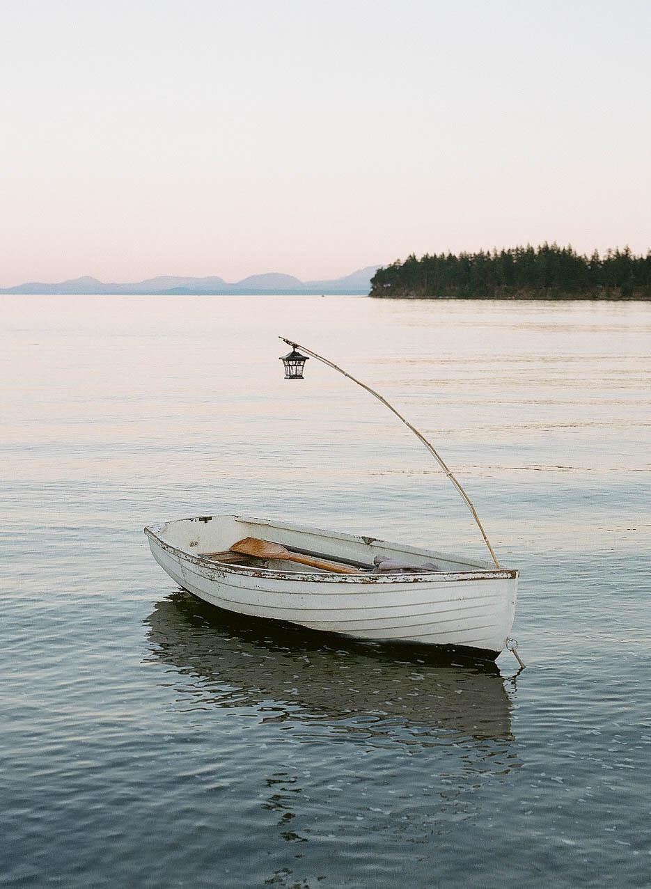 A small white rowboat with oars and a lantern on a pole floats on calm water with an island and mountains in the distance.