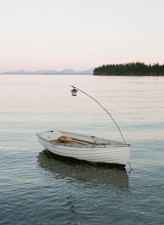 A small white rowboat with oars and a lantern on a pole floats on calm water with an island and mountains in the distance.
