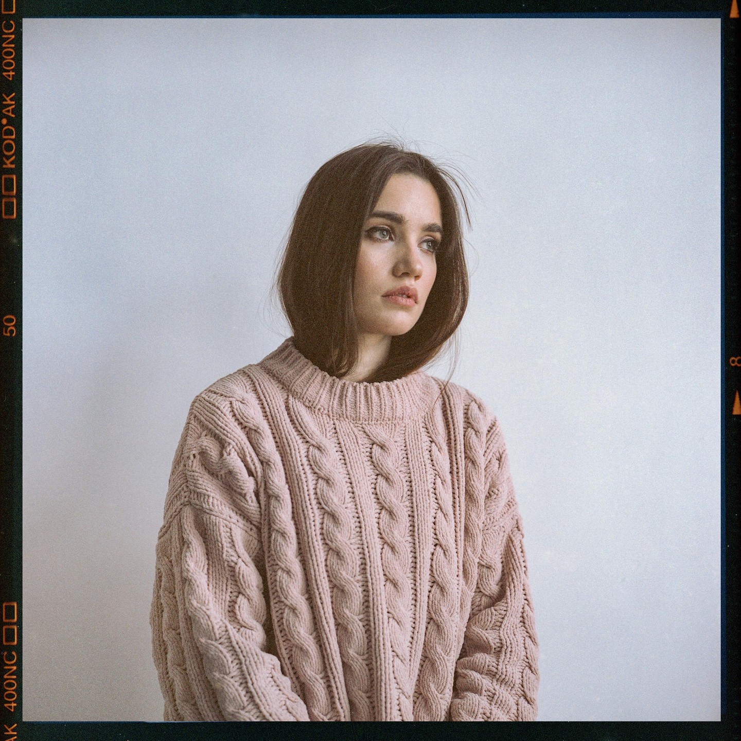 A woman with dark hair wears a pink cable knit sweater and looks to the side.