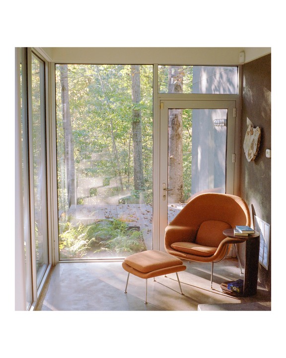 An orange armchair and ottoman sit by a large window looking out onto a forest with sunlight streaming in.