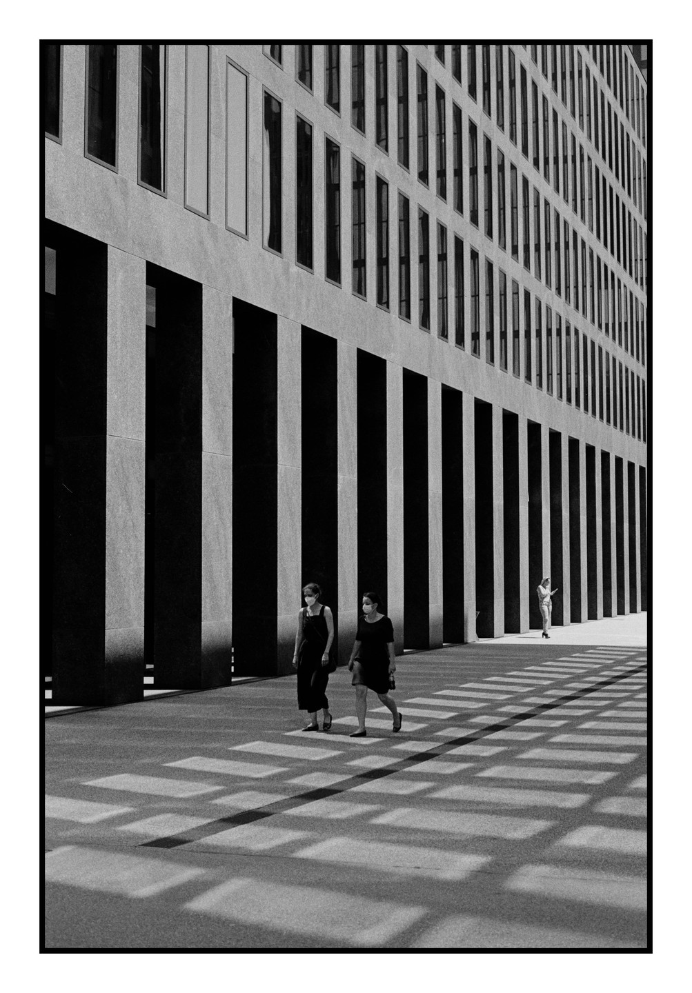 Two women wearing face masks walk past a modern building with strong shadows cast on the ground.