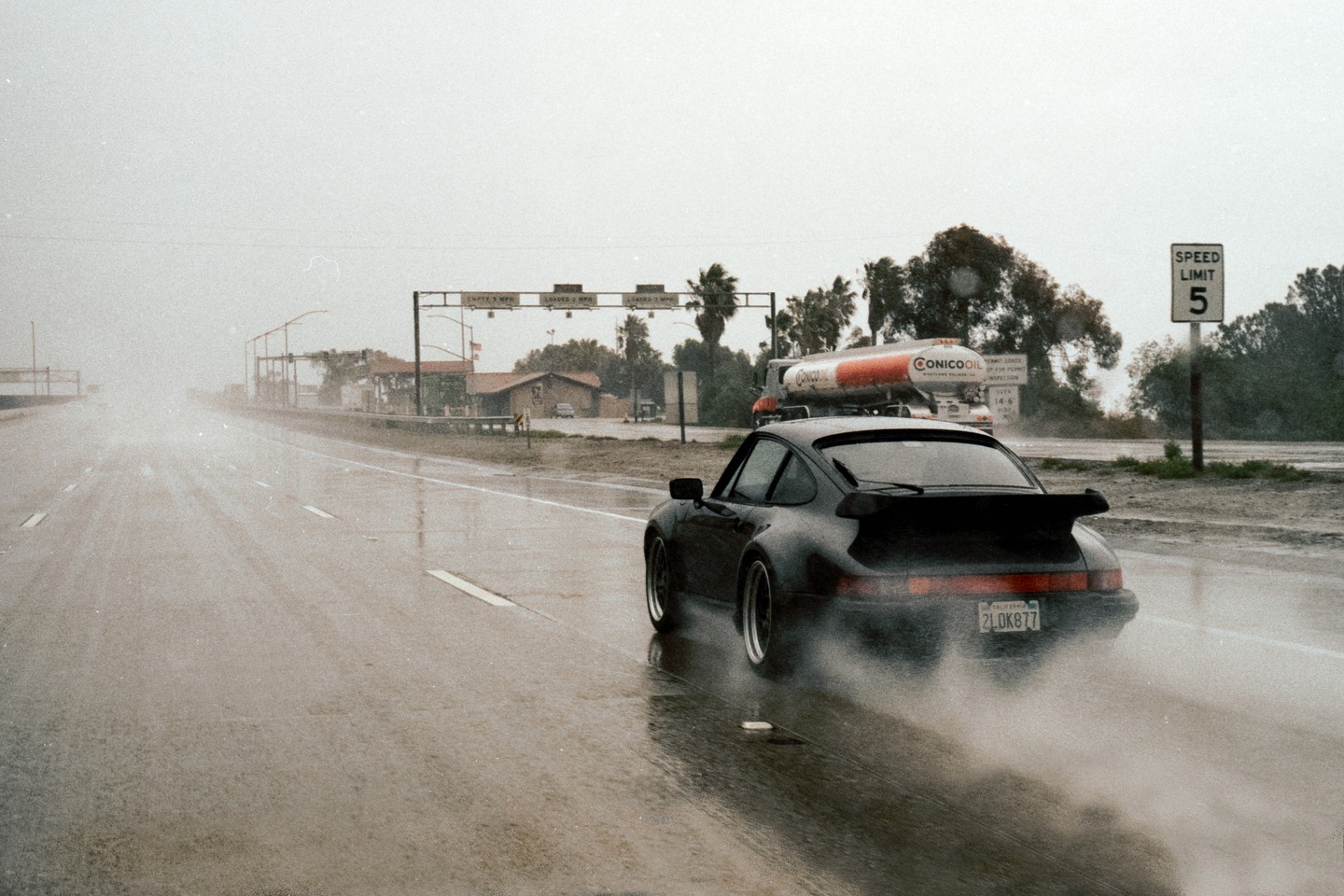 A black car drives on a wet road with a Conoco oil truck in the background.