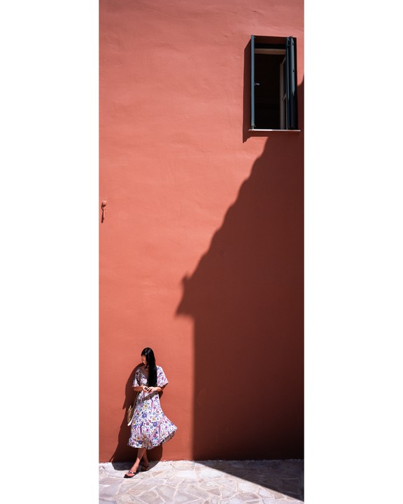 A woman in a floral dress stands against a red wall with a window and a large shadow.