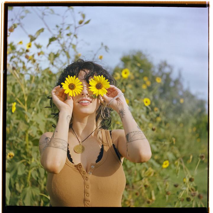 A woman holds two yellow flowers over her eyes in a field of wildflowers.