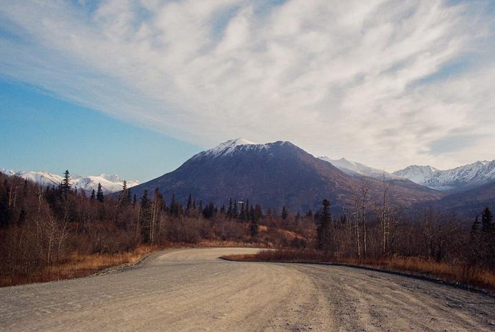 A winding dirt road leads towards a snow-capped mountain range under a partly cloudy sky.